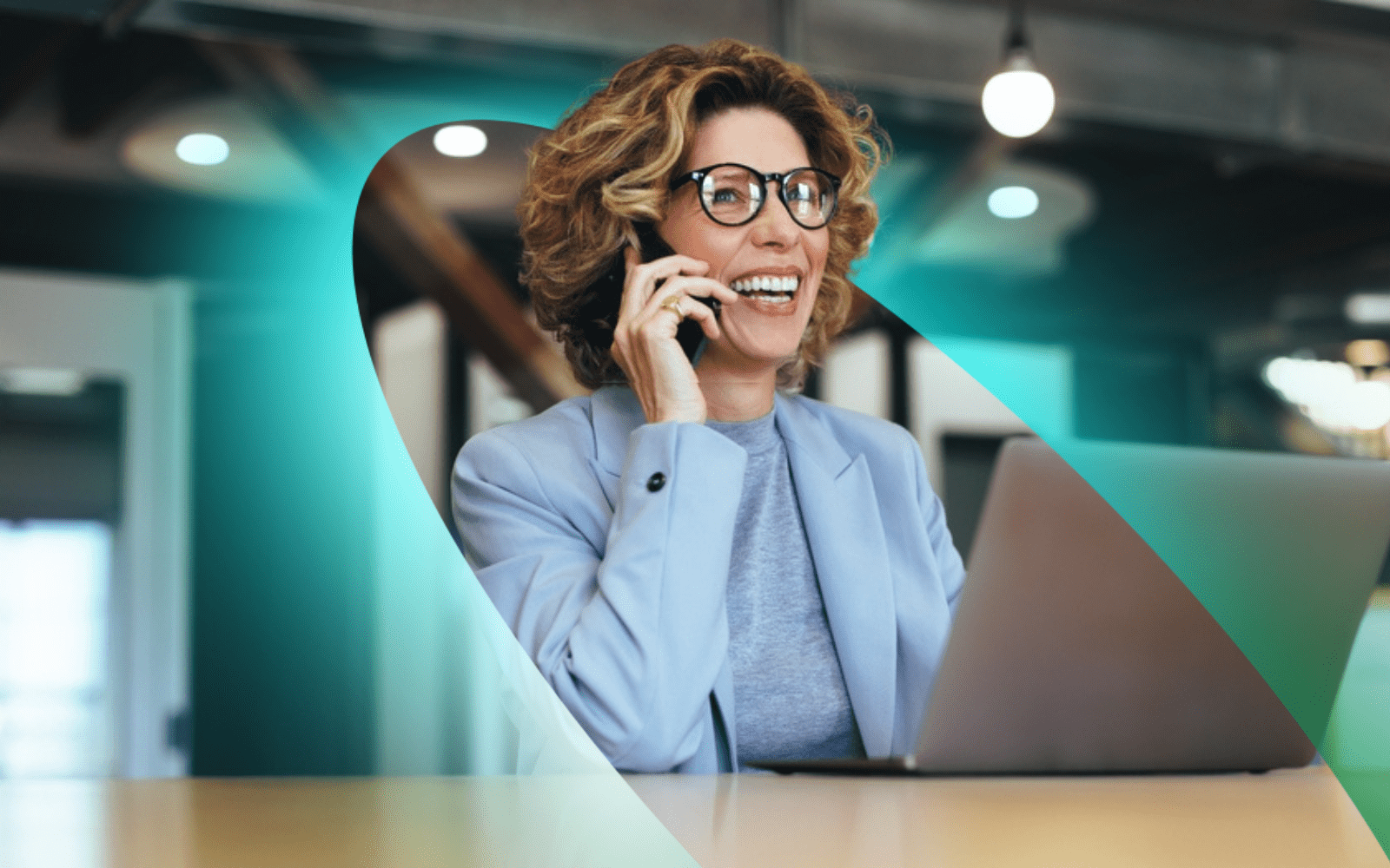 A woman talks happily on a mobile phone with a laptop on the desk in front of her.