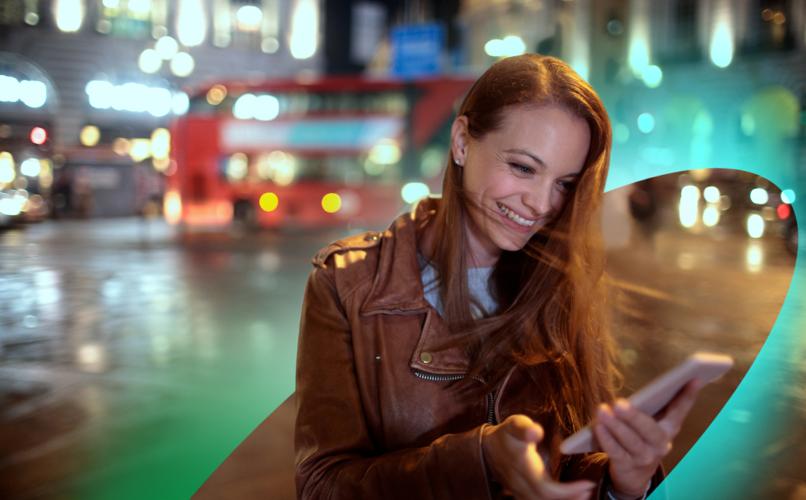 A woman smiling at her mobile phone while standing outside at night time. There is a red London bus behind her.