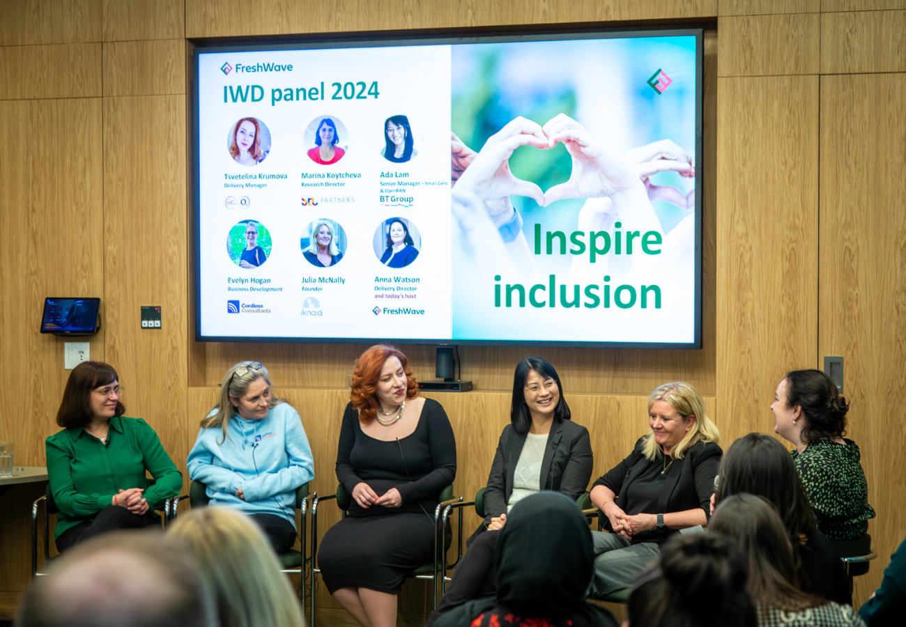 A panel of six women sit on chairs under a large screen that says inspire inclusion. They are celebrating International Women's Day.