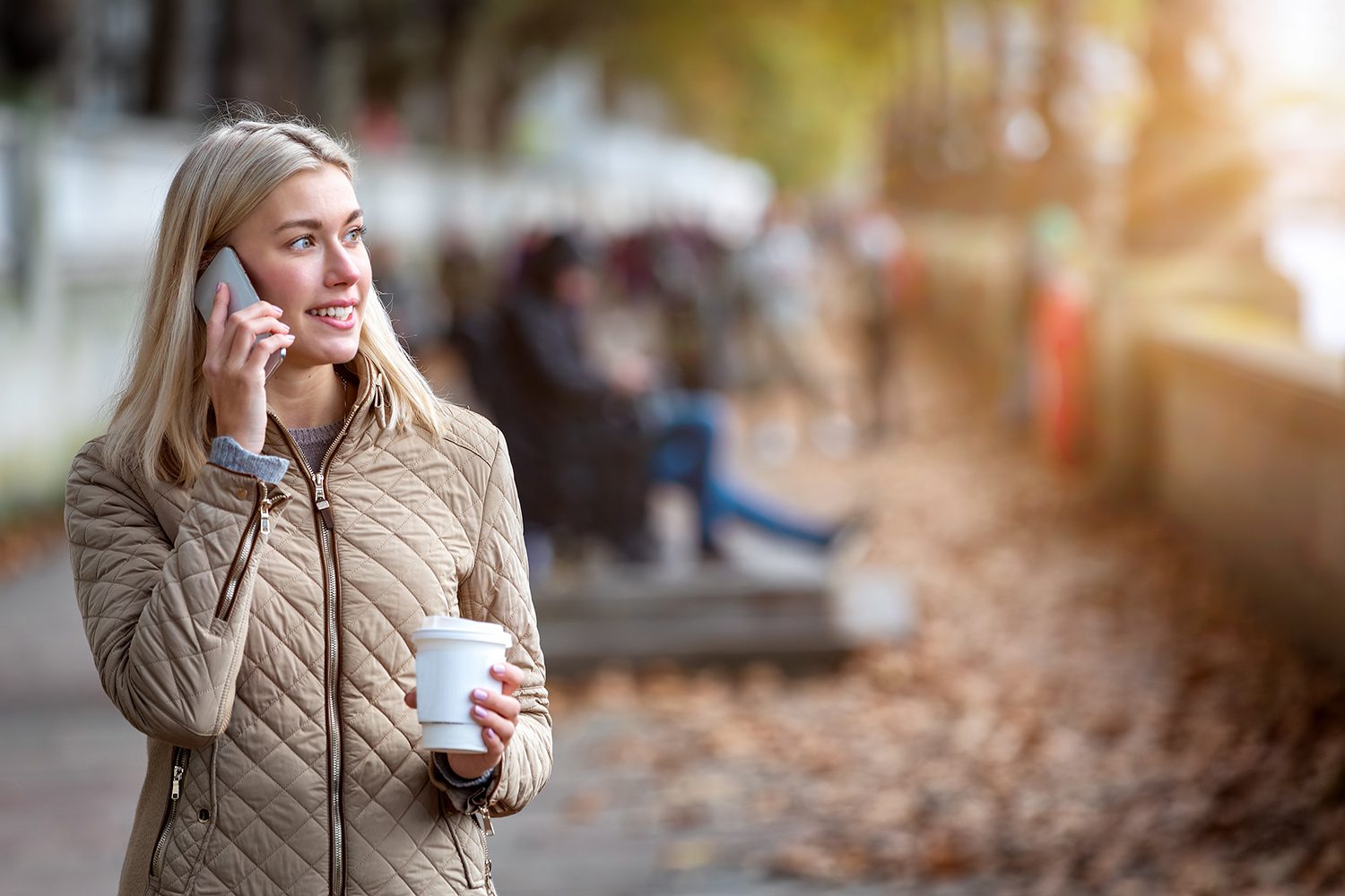 person talking on the phone in a street covered in leaves while holding a coffee cup