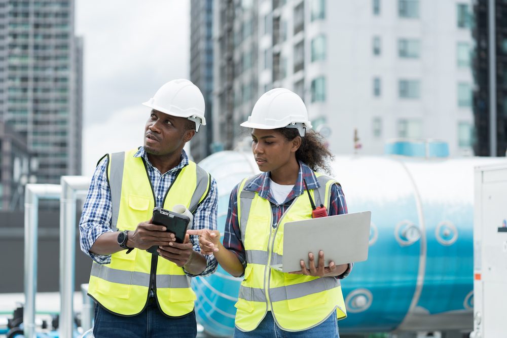 two workers in high visibility jackets and hard hats looking at a laptop and mobile device